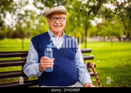 Portrait of happy senior man in park. He is sitting on bench, relaxing and drinking water. Stock Photo