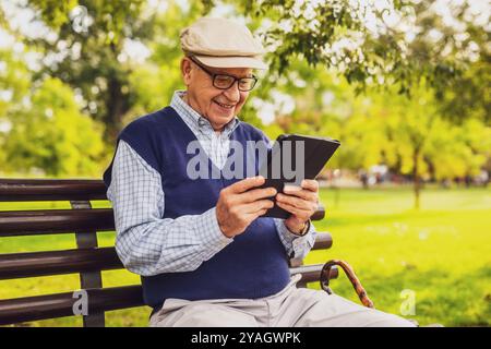 Portrait of happy senior man in park. He is sitting on bench and using digital tablet. Stock Photo