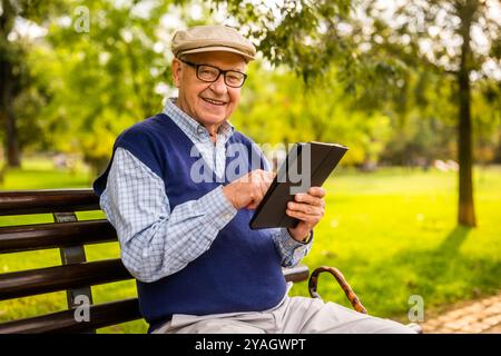 Portrait of happy senior man in park. He is sitting on bench and using digital tablet. Stock Photo