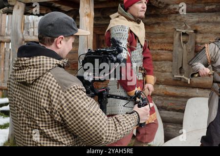 Man wearing medieval costume and holding incidental object while cameraman is filming using professional gear and wooden cabin visible in background Stock Photo
