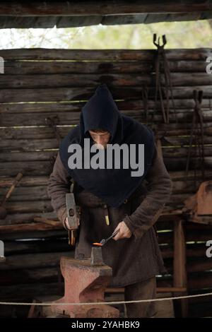 Blacksmith working diligently on metal piece in dimly lit rustic workshop. Various tools hanging on wooden walls in background creating atmosphere of an old-fashioned setting Stock Photo