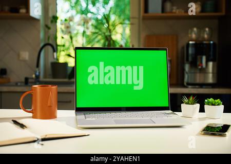 Modern home workspace featuring laptop with green screen, smartphone, coffee mug and small potted plants. Home office for freelancer with kitchen in b Stock Photo