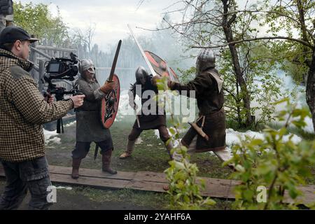 Camera crew capturing medieval battle scene with actors dressed as warriors wielding swords and shields in lush outdoor environment surrounded by trees and bushes Stock Photo