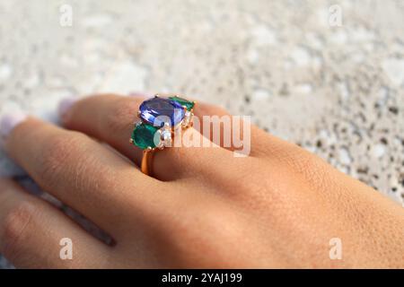hand wearing a gold ring with a large blue gemstone in the center, flanked by two smaller green gemstones and accented with small diamonds, set agains Stock Photo