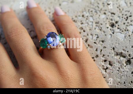 hand wearing a gold ring with a large blue gemstone in the center, flanked by two smaller green gemstones and accented with small diamonds, set agains Stock Photo