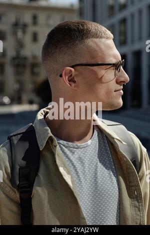 Vertical profile shot of handsome man with tiny septum piercing and glasses standing outside in sunlight while walking in city on nice summer day Stock Photo