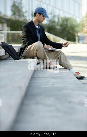 Vertical shot of Asian male software developer writing code on laptop looking at computer screen while sitting on concrete stairs in city street, copy space Stock Photo