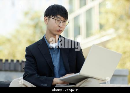 Medium shot of concentrated Asian male businessman in black jacket looking through thin framed glasses at laptop screen while working remotely from sunlit city street, copy space Stock Photo