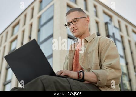 Low angle shot of handsome male software engineer wearing glasses writing code developing applications on laptop while sitting outside in front of business building in city street, copy space Stock Photo