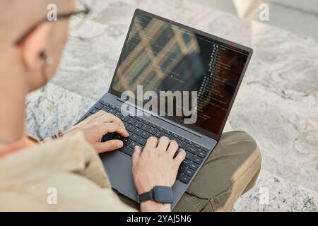 Over shoulder shot of unrecognizable male IT programmer writing code on laptop typing on computer keyboard while sitting on concrete steps outside in city street, copy space Stock Photo