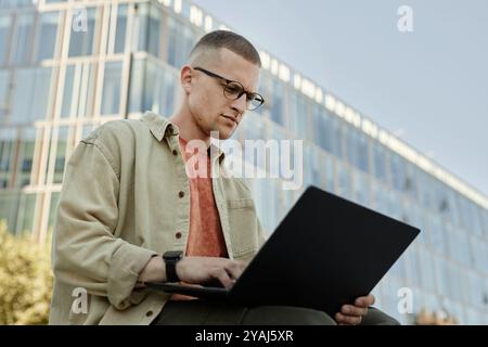 Medium shot of serious male IT programmer with glasses and tiny septum using laptop working outdoors while sitting outside in city street, copy space Stock Photo