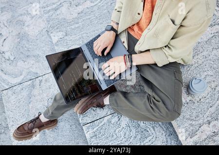 High angle of unrecognizable male IT developer using laptop to write code sitting on concrete steps with reusable cup placed nearby while working outside in city, copy space Stock Photo