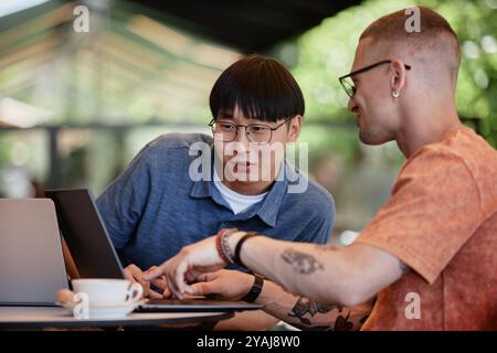 Medium shot of Asian male IT developer looking at laptop screen while collaborating with colleague at work using computers to write code at cafe table, copy space Stock Photo