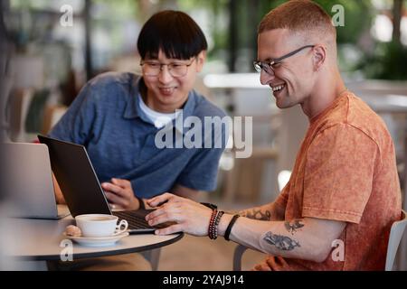 Side view of smiling male software engineer typing on laptop writing code while having fun working collaboratively with colleague during meeting over coffee in outdoor seating area of cafe, copy space Stock Photo