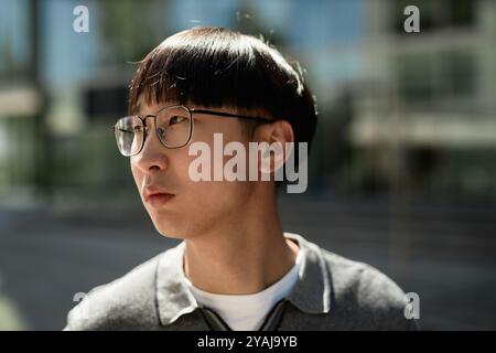 Close up of adult Asian man with glasses looking away lost in thought while standing outside in sunlight on blurred background, copy space Stock Photo