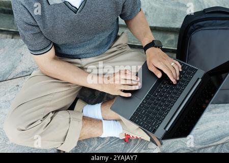 Top view of unrecognizable businessman typing on laptop keyboard while sitting on pavement working remotely outside in city street, copy space Stock Photo