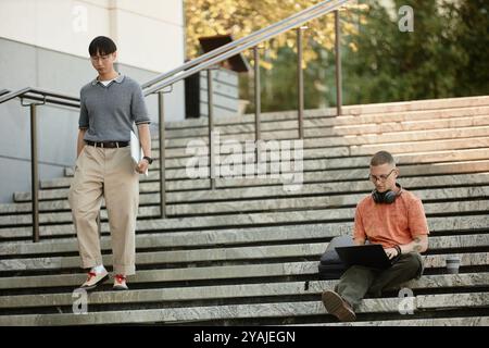 Wide angle shot of Asian businessman in office clothing walking down stairs where male colleague using laptop to work sitting on steps outside in public space in city, copy space Stock Photo
