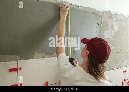 Woman tiling bathroom walls, using tape measure for measuring length. Process of installation of tiles in the bathroom step by step. DIY home improvem Stock Photo