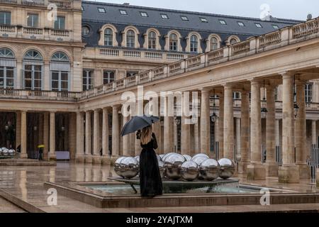A blonde woman dressed in black with a black umbrella outside the ancient Palais-Royal in Paris, France Stock Photo
