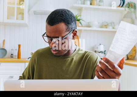 Young focused african american man wearing glasses working remotely from home sitting in the kitchen and checking bills, managing monthly expenses and finances Stock Photo