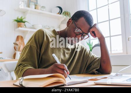 Young black man studying at home, focused on taking notes from a textbook, using a laptop for resources, surrounded by educational materials and natural light Stock Photo