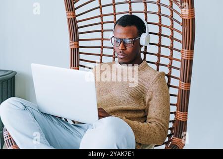 Young professional is working remotely, sitting in a comfortable hanging chair while typing on his laptop and wearing headphones Stock Photo