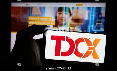Person holding mobile phone with logo of Singaporean customer experience company TDCX on screen in front of web page. Focus on phone display. Stock Photo