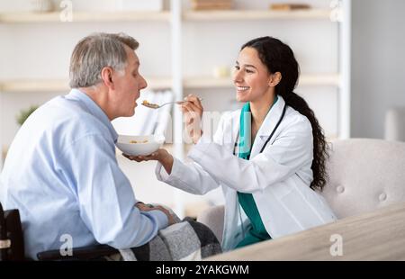 Medical assistance for elderly people with disabilities. Nurse feeding senior handicapped man in wheelchair at home Stock Photo