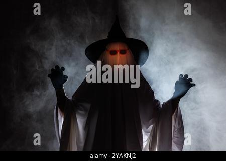 Man in scary ghost costume for Halloween and witch hat on dark mystical background in smoke, Concept of Halloween Stock Photo