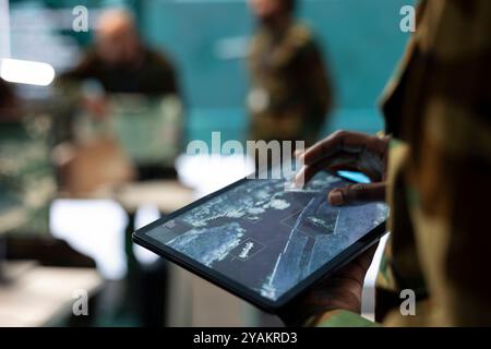 Recruit analyzing deployed infantry unit ongoing operation on his tablet, using surveillance satellite system and a world map. IT engineer working with high tech gear for reconnaissance. Close up. Stock Photo