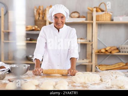 Smiling mature female baker rolling out dough in bakery Stock Photo