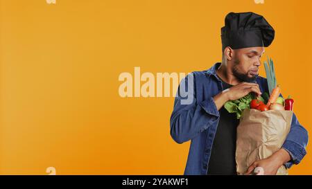African american cook examining his groceries in a paper bag before preparing a vegan meal, feeling pleased about cooking food with ethically sourced ingredients. Sustainable lifestyle. Camera B. Stock Photo