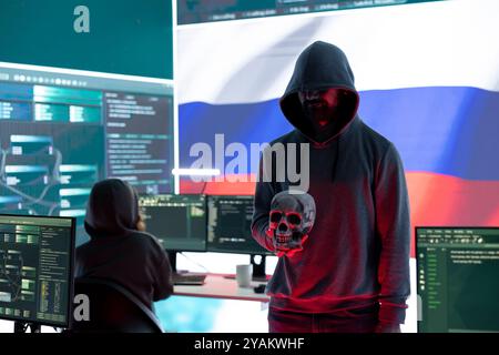 Conceptual image of a hooded russian hacker creating hazardous alerts, holds a skull and warning of death threats. Russian federation works on cybercrime and violence, panic and torment. Stock Photo