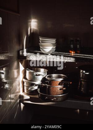 A dimly lit kitchen corner with dramatic sunlight streaming in, illuminating stacked dishes, bowls, pots and pans. Stock Photo