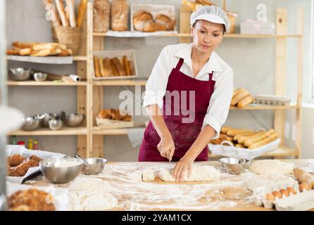 Focused female baker in apron working at bakery, cutting dough into pieces Stock Photo