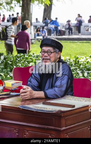 HANOI, VIETNAM, NOVEMBER 14: Old vietnamese man in national dress writes hieroglyphs on old paper at the street at November 13, 2016 in Hanoi, Vietnam Stock Photo