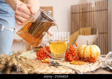 Female hand pouring tea from teapot into cup near bunch of wheat and rowan on table in room. Closeup Stock Photo