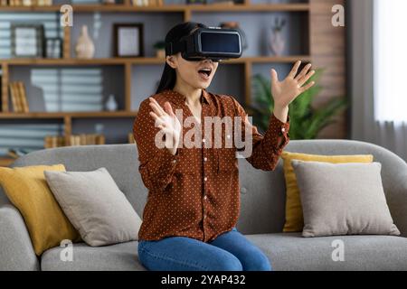 Excited asian woman wearing virtual reality headset experiencing immersive digital content in cozy living room. Expression shows amazement and engagement with modern VR technology and lifestyle. Stock Photo