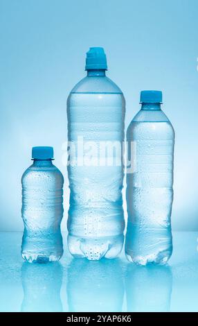 Three plastic bottles with frosted caps filled with water of different sizes on wet reflective blue surface against blue background Stock Photo