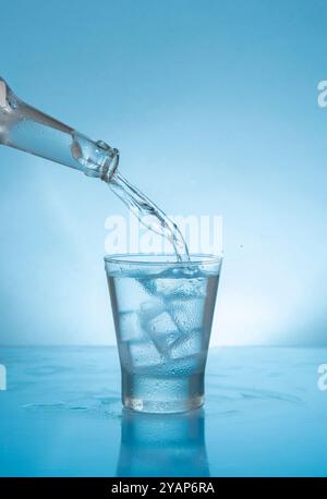 Glass bottle filling with water a glass glass with ice on wet reflective blue surface against blue background Stock Photo