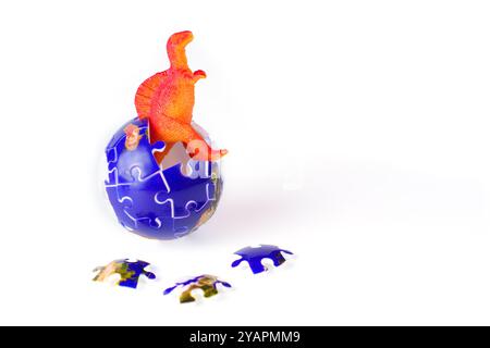 Orange dinosaur hatching from a puzzle globe with scattered pieces, set against a white background, symbolizing the rise and fall of species and the i Stock Photo
