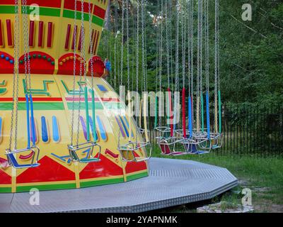 Empty old-fashioned merry-go-round in a city park. Red and blue wooden seats on metal chains of swing carousel. Trees on the background, fallen leaves Stock Photo