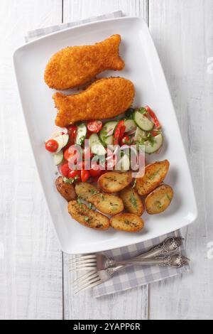 Breaded fried hake fish with fresh salad and baked potato wedges close-up in a plate on a wooden table. Vertical top view from above Stock Photo