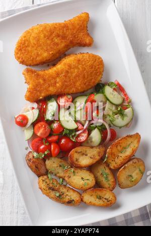 Breaded fried fish with fresh salad and country potatoes close-up in a plate on a wooden table. Vertical top view from above Stock Photo