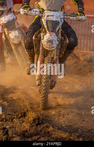 Front view of an off-road motocross bike riding on muddy terrain with the wheels, boots and bike covered in mud escaping from a pursuer in an enduro e Stock Photo