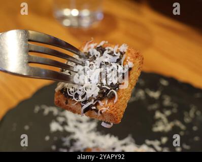 A fork is holding a piece of food with coconut flakes on it. The food is on a black plate Stock Photo