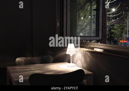 A table with a lamp on it and a chair next to it. The lamp is lit up and the room is dark Stock Photo