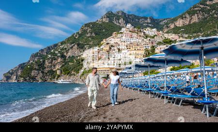 A couple enjoys a romantic walk along the pebbled Amalfi Coast beach, surrounded by colorful houses on a hillside. The sky is clear and blue, perfect for an unforgettable day by the sea. Stock Photo