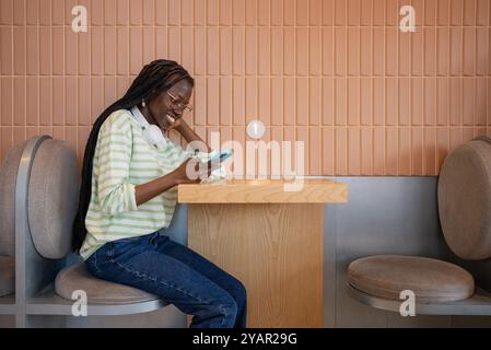 Young woman in a cozy cafe, smiling and using her smartphone, enjoying a break with wireless earphones, showcasing a carefree digital lifestyle Stock Photo