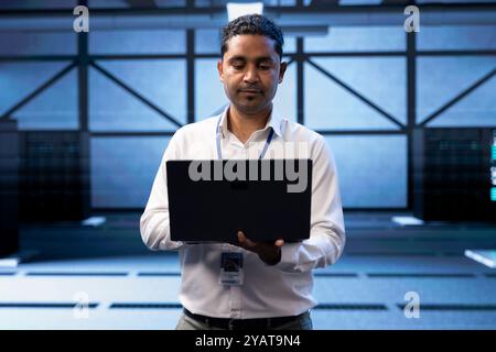 Engineer in server farm checking recovery plan on laptop, monitoring data center energy consumption across components, making sure sensors are functioning optimally, doing maintenance on equipment Stock Photo